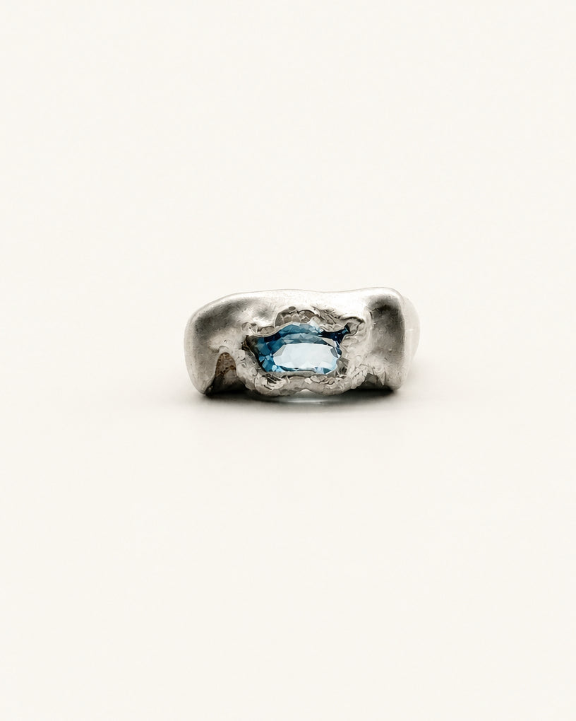 An abstract, chunky sterling silver ring featuring a central oval-cut light blue gemstone, photographed on a clean, solid off-white background.
